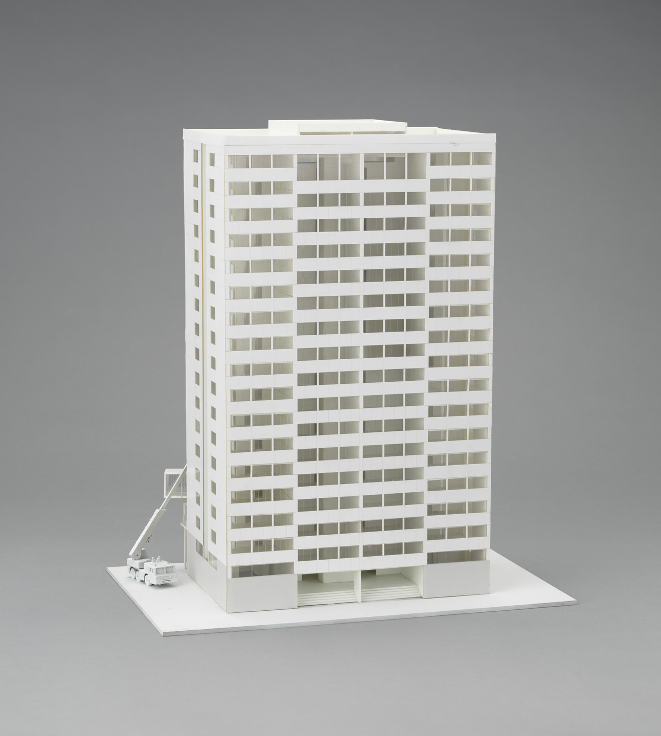 A model of a tall, white, rectangular building with many identical windows is centered on a gray background. A small crane and truck are positioned to the left of the building. This is an AI generated description. To give feedback, email ai@moma.org.