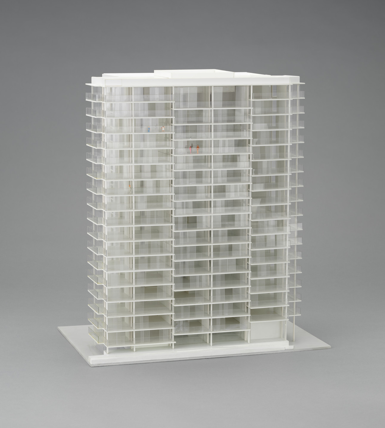 The image shows a white architectural model of a high-rise building with multiple floors and balconies, viewed against a gray background. Small, colored figurines are placed on some of the balconies. This is an AI generated description. To give feedback, email ai@moma.org.