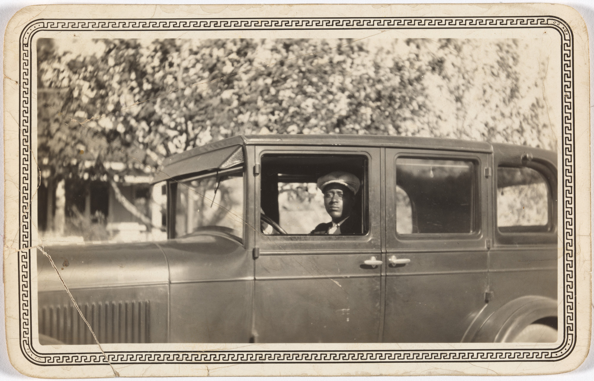 A vintage photograph shows a person in a dark car, wearing a light-colored cap, with trees and buildings slightly blurred in the background. The photo is bordered with a decorative pattern. This is an AI generated description. To give feedback, email ai@moma.org.