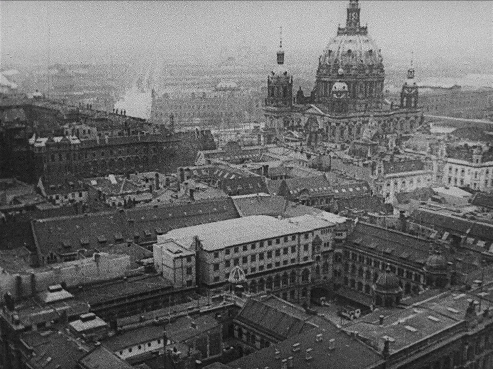 A sepia-toned image depicts an aerial view of a densely built cityscape dominated by an ornate domed cathedral surrounded by numerous tightly packed buildings and rooftops. The background is hazy, suggesting distance or fog. This is an AI generated description. To give feedback, email ai@moma.org.