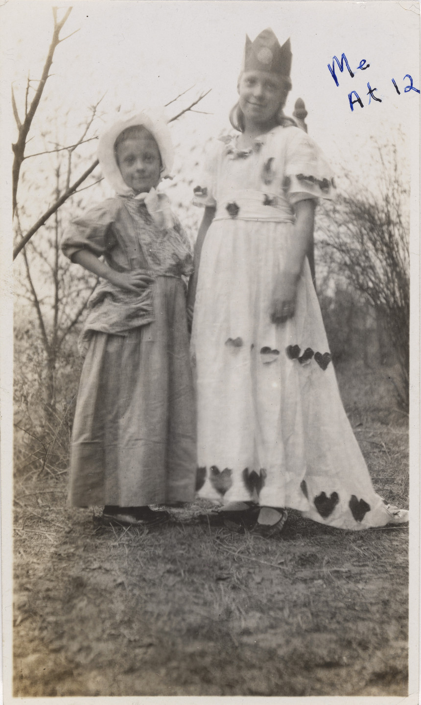 Two children are standing on the ground near bare trees, one in a bonnet and dress, and the other in a long white dress with heart shapes and a crown. The photo is in black and white. This is an AI generated description. To give feedback, email ai@moma.org.