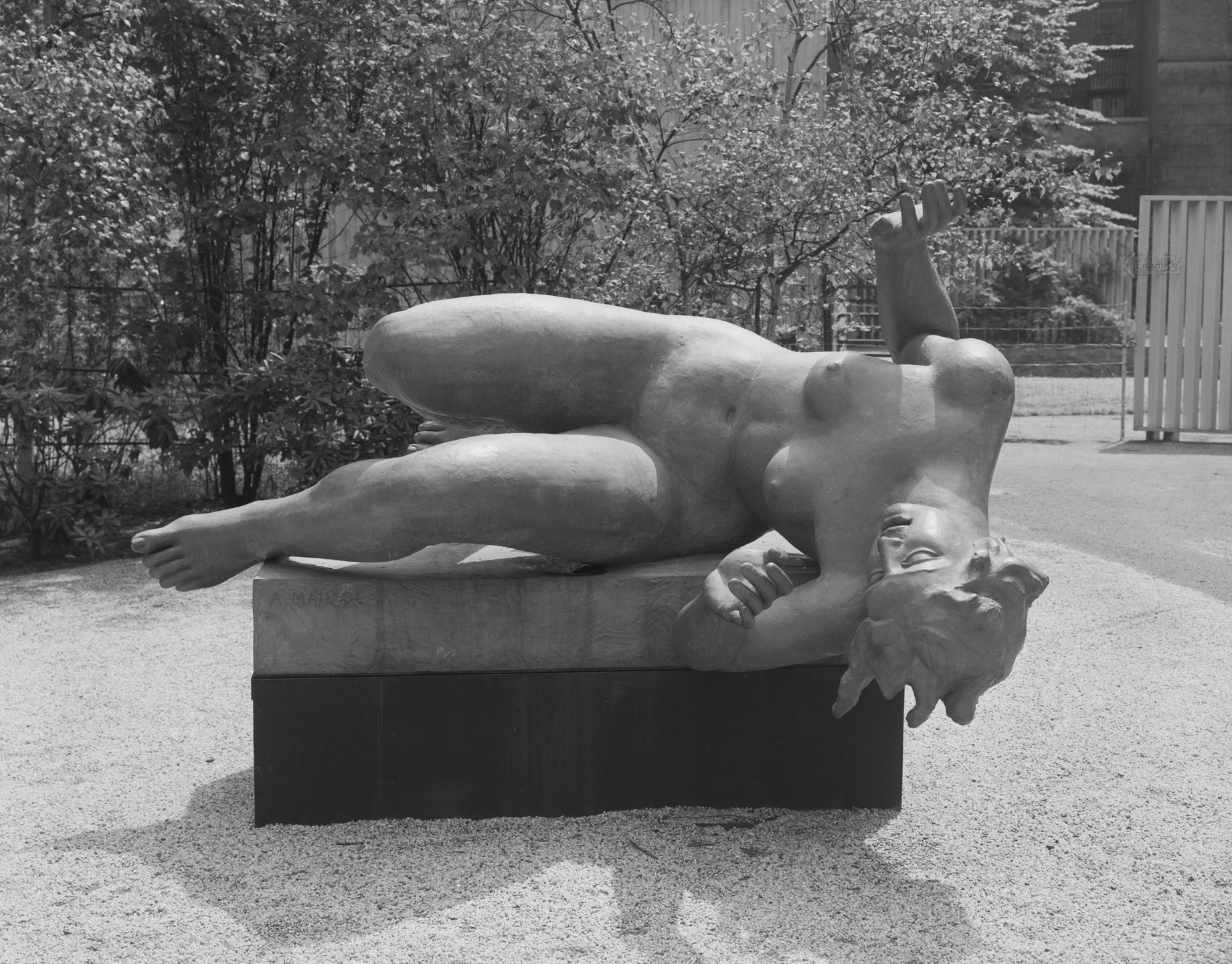 A stone sculpture of a reclining human figure rests on a pedestal, surrounded by gravel and trees in the background. The figure's head and legs extend towards the viewer, and one arm is bent with an open hand. This is an AI generated description. To give feedback, email ai@moma.org.