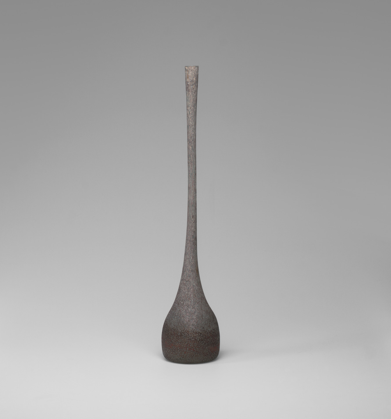 A tall, thin vase with a rounded base tapering to a narrow neck stands against a gray background. The vase is predominantly dark gray with subtle speckles of brown. This is an AI generated description. To give feedback, email ai@moma.org.