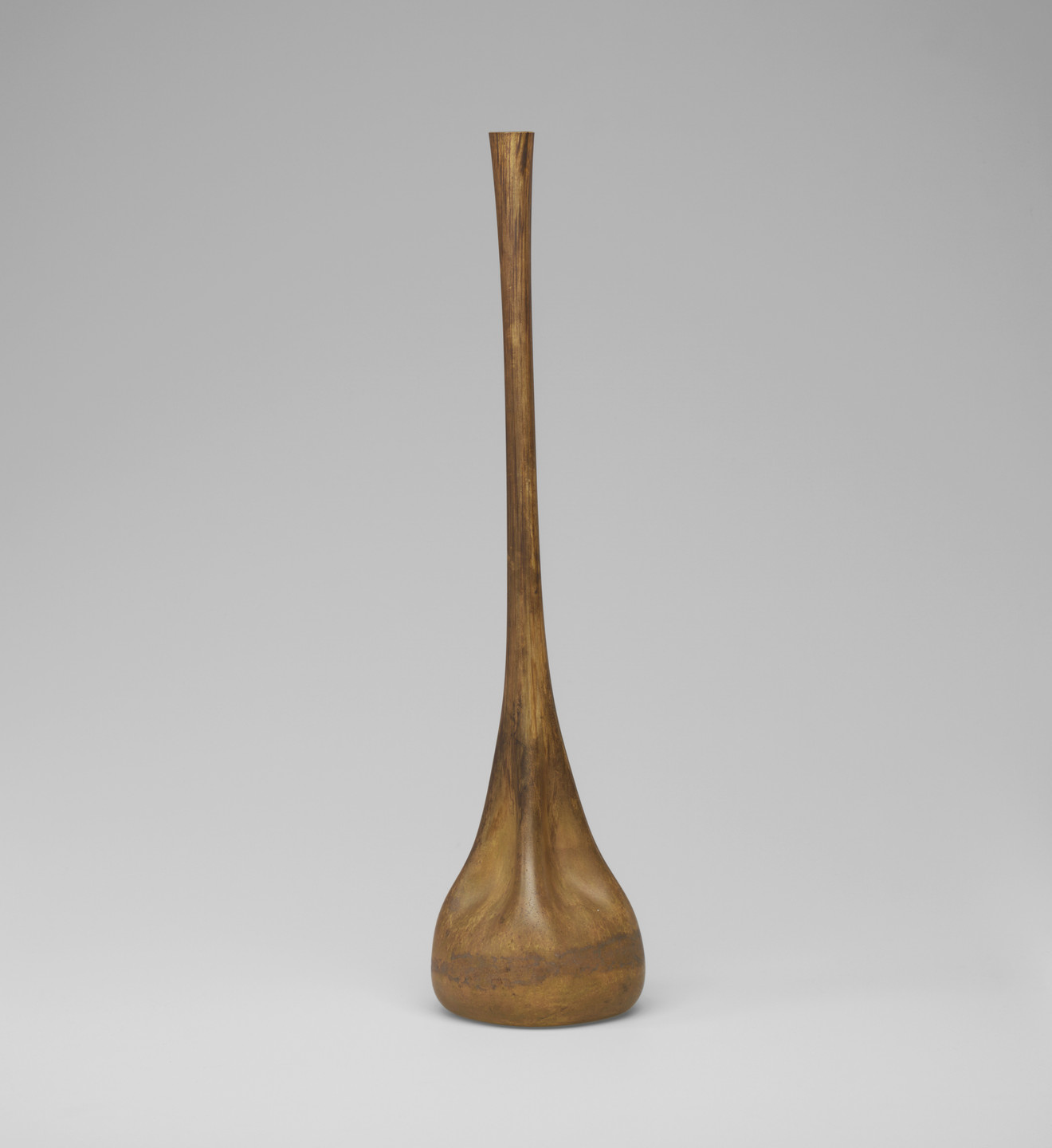 A tall, slender handpainted glass vase with a wide base tapers into a narrow neck. The vase is a dark mustard gold color and sits against a plain, light grey background. This is an AI generated description. To give feedback, email ai@moma.org.
