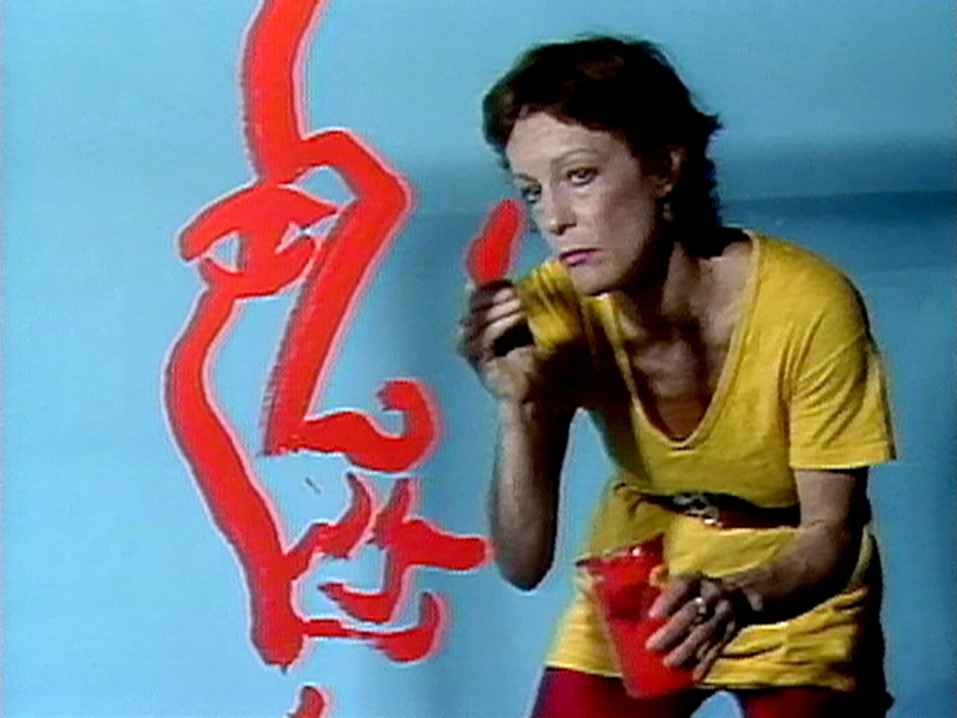 A person in a yellow outfit holds a red paintbrush and container, painting a large, red face outline on a light blue wall. The person looks toward the camera while painting. This is an AI generated description. To give feedback, email ai@moma.org.