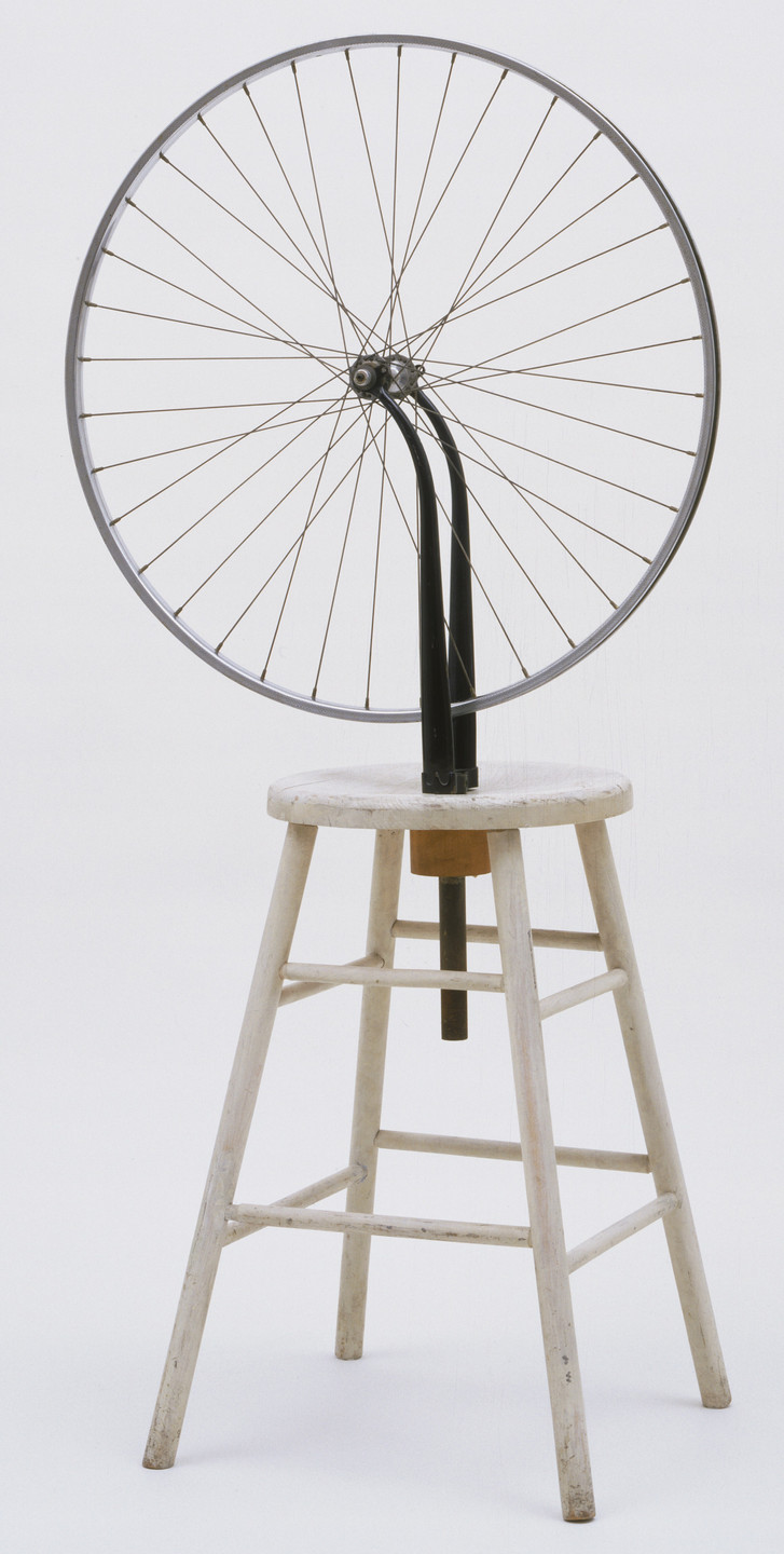 A bicycle wheel (without a tire) is fixed upside down on a plain white wooden stool with a black fork supporting the wheel. The background is completely white. This is an AI generated description. To give feedback, email ai@moma.org.