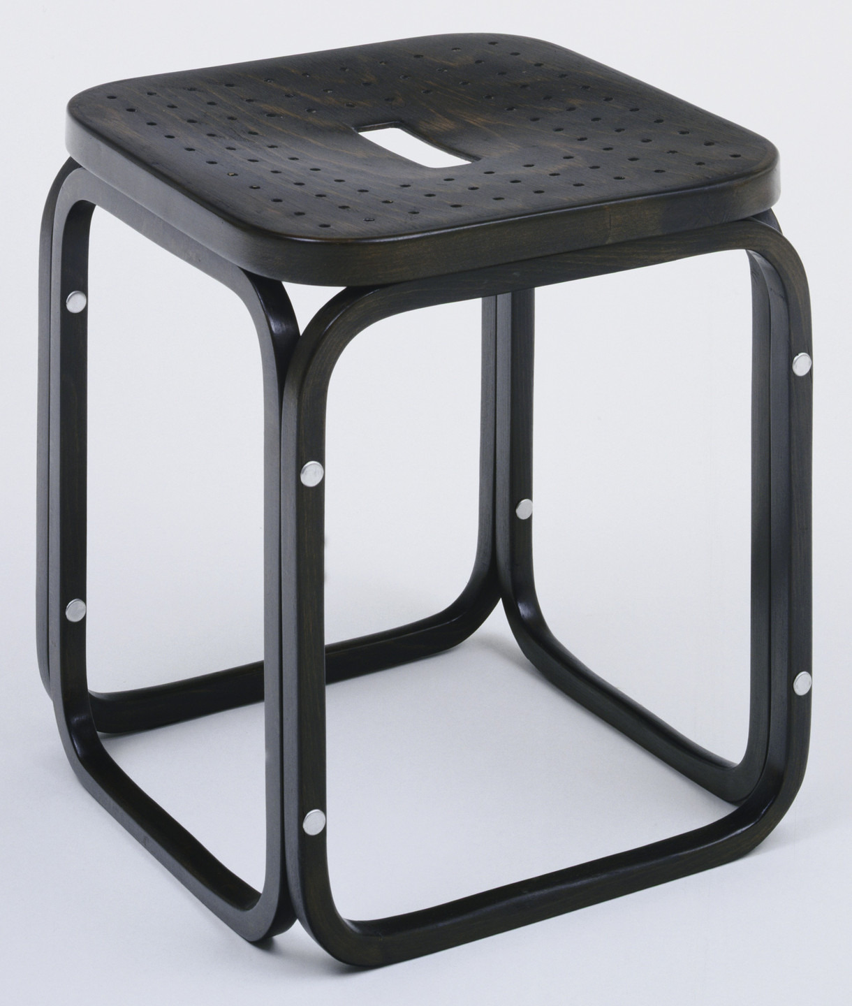 A dark brown wooden stool with a square perforated seat and a rectangular hole in its center. The stool has a minimalist design with rounded corners and silver rivets at the joint points. This is an AI generated description. To give feedback, email ai@moma.org.