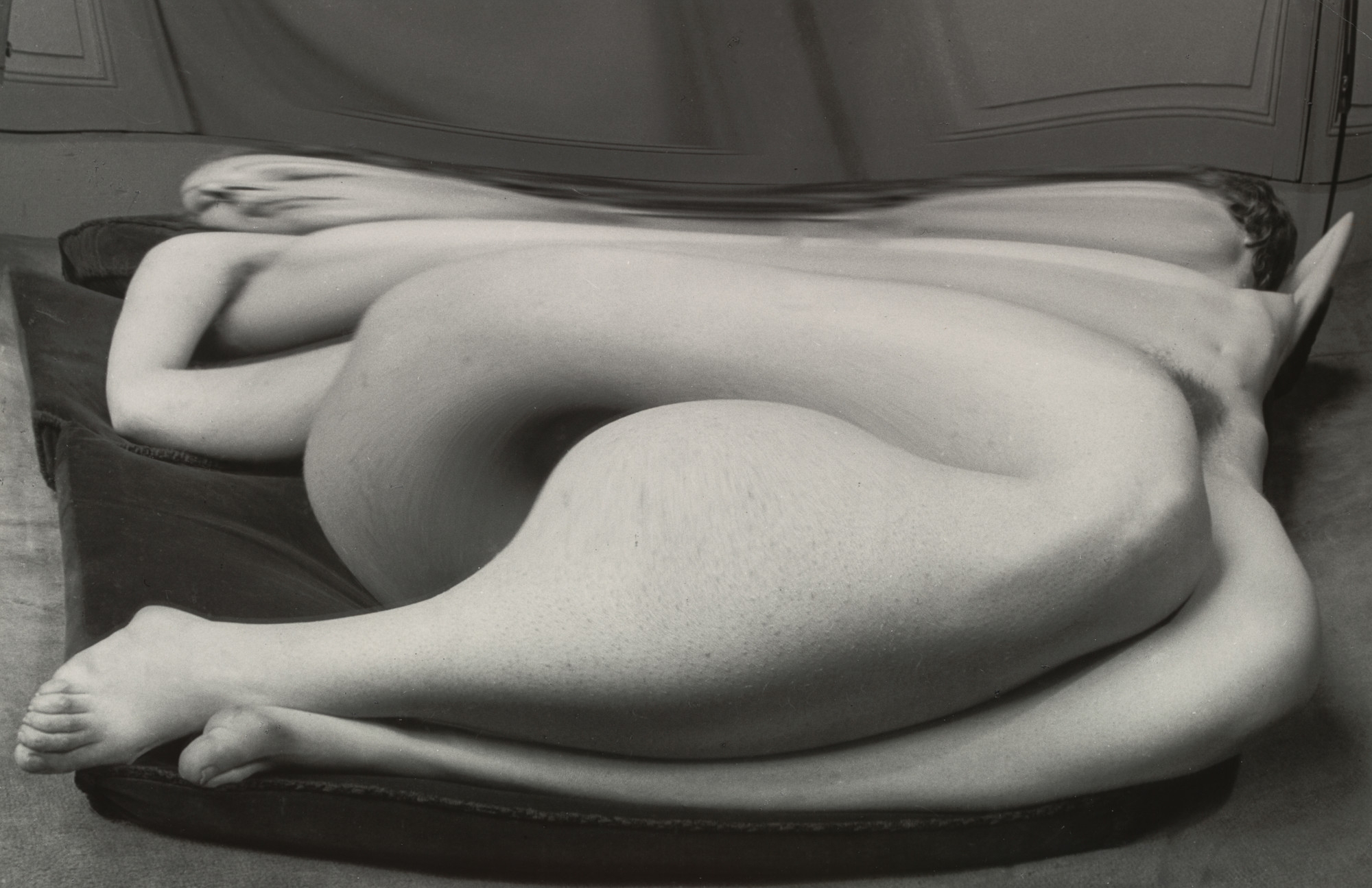 The grayscale image depicts a distorted human figure lying on a dark surface. The body appears warped and stretched, with prominent curvature and overlapping limbs. This is an AI generated description. To give feedback, email ai@moma.org.