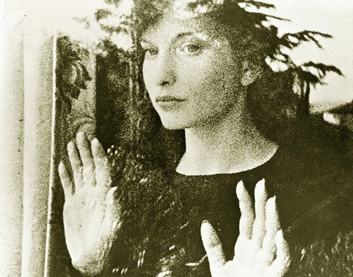 A person with dark hair stands behind a glass window, facing forward with hands pressed against the glass. The image is in black and white with visible reflections and foliage in the background. This is an AI generated description. To give feedback, email ai@moma.org.