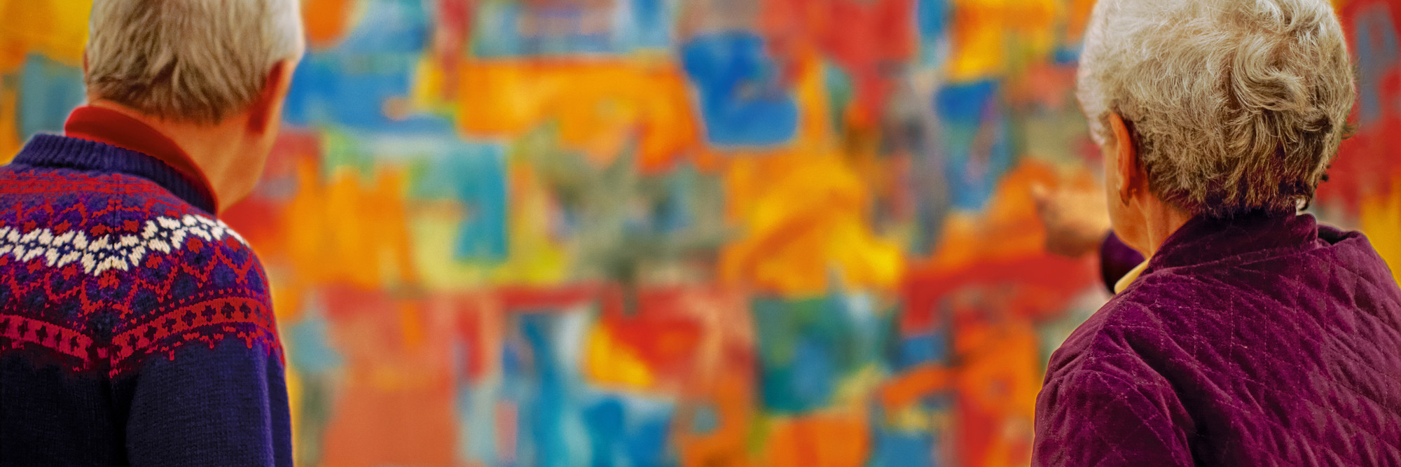 Photo: Jason Brownrigg. Digital image © 2023 The Museum of Modern Art, New York Image description: A photograph of two adults on either side of the frame looking at the out-of-focus, large, and colorful painting Map by the artist Jasper Johns.