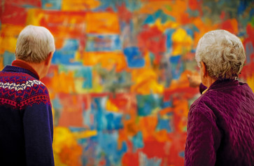 Photo: Jason Brownrigg. Digital image © 2023 The Museum of Modern Art, New York Image description: A photograph of two adults on either side of the frame looking at the out-of-focus, large, and colorful painting Map by the artist Jasper Johns.