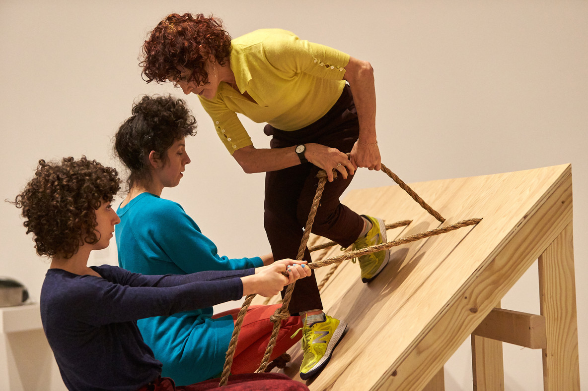 Slant Board features a 45-degree wooden incline with ropes fastened along the top. Left to right, Talya Epstein, Lee Relvas, and Jody Oberfelder brace themselves with the ropes and move across the board, navigating one another’s presence. Occasionally, the performers rest, standing or crouching while bracing themselves with the ropes.