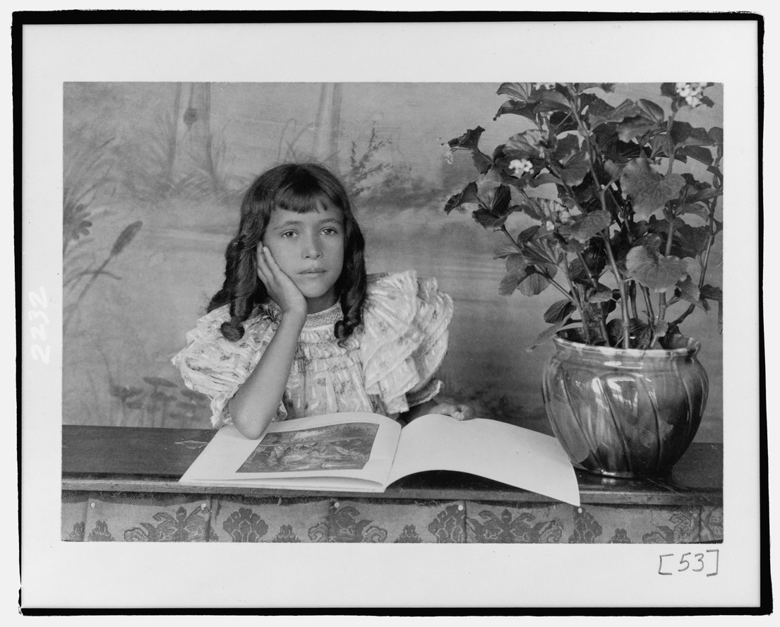 A photo featured in Wayward Lives, Beautiful Experiments: Thomas E. Askew. African American girl, half-length portrait, with the right hand to cheek, with illustrated book on table. c. 1900
