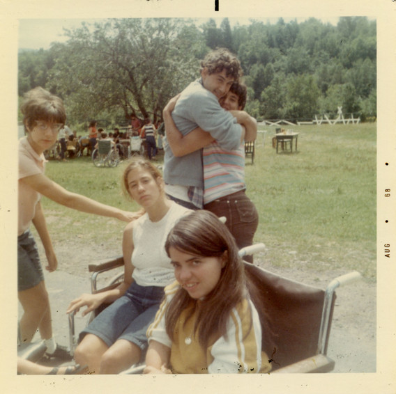 Crip Camp. 2019. USA. Directed by Nicole Newnham, Jim LeBrecht. Image description: Several young adults gather in a grassy outdoor space. In the foreground, two people seated in wheelchairs look directly at the camera. A third stands to the left, one outstretched hand touching the back of one of the wheelchairs. Behind these three, two more young people stand in a tight embrace, grinning. In the distance, a larger group of people is gathered together below a tree.