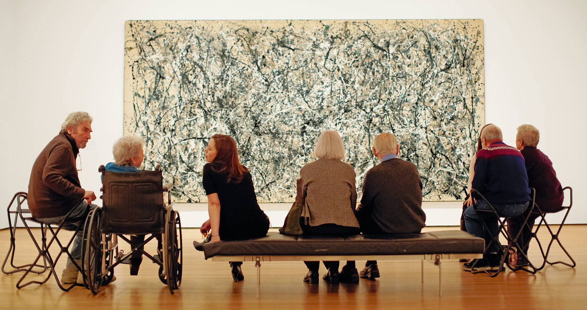 Older adults take in a Jackson Pollock during a MoMA Access Program. From “For Caregivers: A Guide for Connecting Creatively.” Image description: In a gallery, a group of seven adults sits with their backs to the camera, in front of a large abstract painting. Most of the adults have gray hair. One adult with long light brown hair is turned to the left, speaking to a gray-haired individual in a wheelchair. In the background, the painting, so large it is only partially visible, features arcing spatters of black, white, and gray paint.