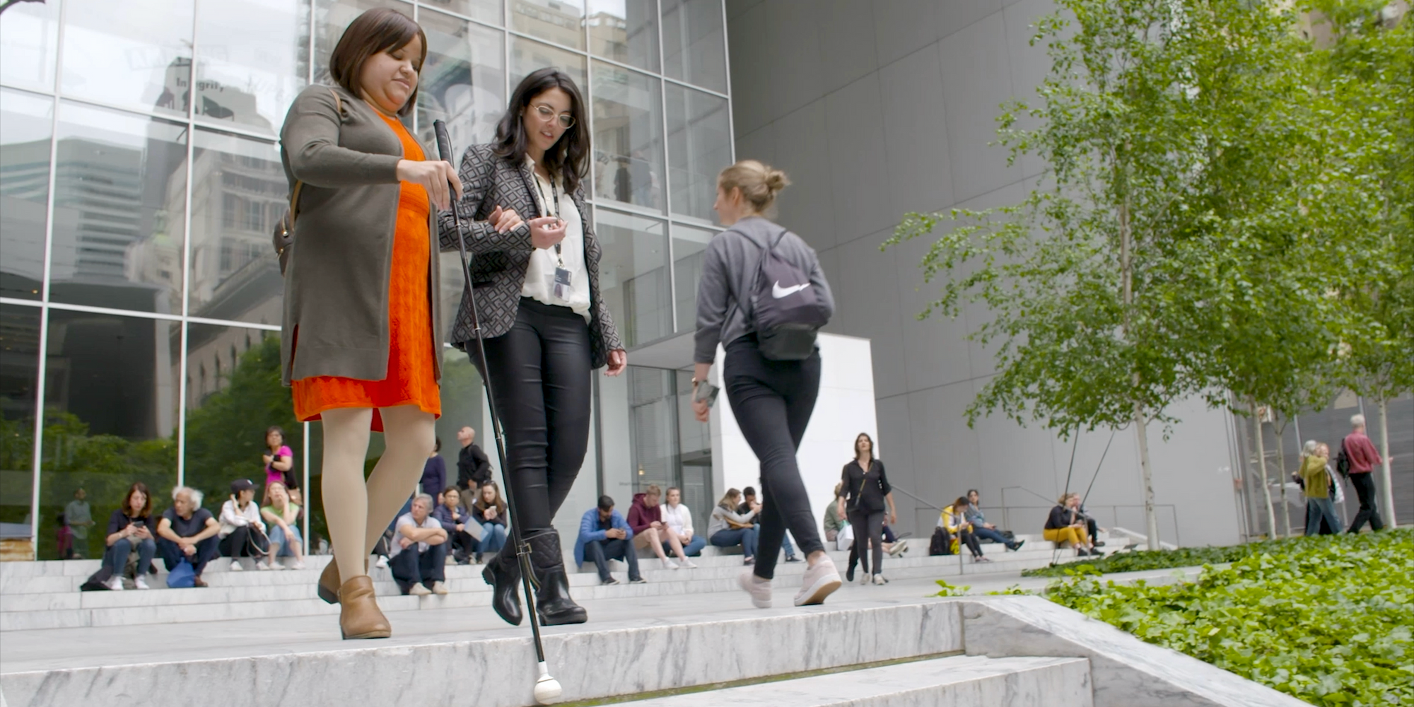 Nefertiti Matos walks with a sighted guide in MoMA’s Sculpture Garden. Video still image by J6 MediaWorks, from Disability Equality and Museums Episode 7: Meet Nefertiti. Image description: Two women walk together down the steps of MoMA’s sculpture garden, with their arms linked. The woman to the left wears an orange dress and extends a long black cane in front of her. The other woman wears black pants and boots. Both women look downward.