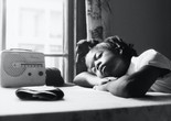 Garrett Bradley. America. 2019. Multichannel video installation. Courtesy the artist Caption: A black-and-white image of a young Black schoolgirl resting in the sunlight from a bright window with an old-time radio. Alt-text: Garrett Bradley. America. 2019. Multichannel video installation. Courtesy the artist. Image Description: A young Black schoolgirl sits at an apartment table draped with a tablecloth. In the black-and-white image, her arms are folded atop one another, the crook of her elbow making a cradle for her lowered head. Her hair is pulled into a half-up, half-down style with a big bow adorning the top of her head. Her face, in peaceful rest, is lit by a bright window. An old-time radio sits on the table next to her.