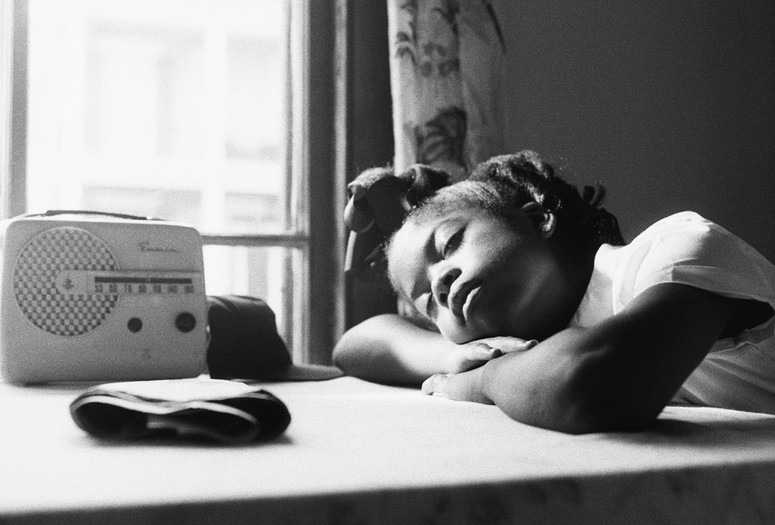 Garrett Bradley. America. 2019. Multichannel video installation. Courtesy the artist Caption: A black-and-white image of a young Black schoolgirl resting in the sunlight from a bright window with an old-time radio. Alt-text: Garrett Bradley. America. 2019. Multichannel video installation. Courtesy the artist. Image Description: A young Black schoolgirl sits at an apartment table draped with a tablecloth. In the black-and-white image, her arms are folded atop one another, the crook of her elbow making a cradle for her lowered head. Her hair is pulled into a half-up, half-down style with a big bow adorning the top of her head. Her face, in peaceful rest, is lit by a bright window. An old-time radio sits on the table next to her.