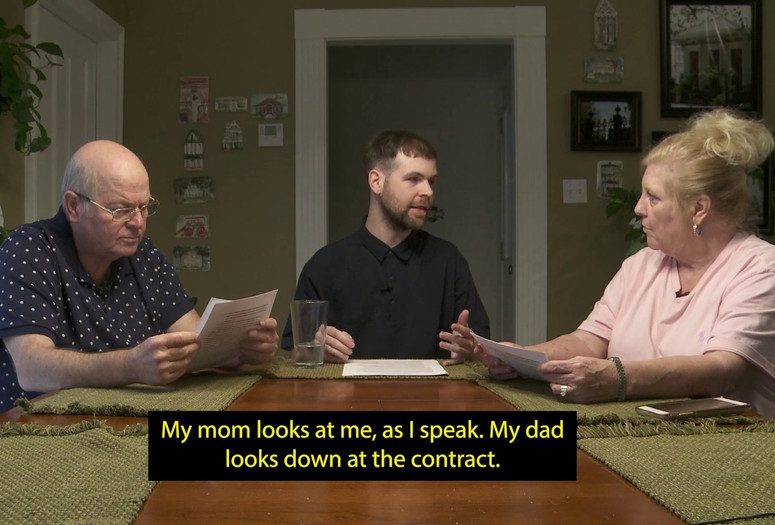 Shared Resources. 2021. Directed by Jordan Lord. Courtesy the filmmaker. Image description: Three white people sit at a table together, each with a set of white papers in front of them. The figure on the far left is a bald man in his 60s, who reads the papers intently. The figure in the center is a person in their late 20s, who looks to a woman in her 60s with blonde hair, who holds the papers in her hands and looks back at them. A caption appears over the image that reads: “My mom looks at me, as I speak. My dad looks down at the contract.”