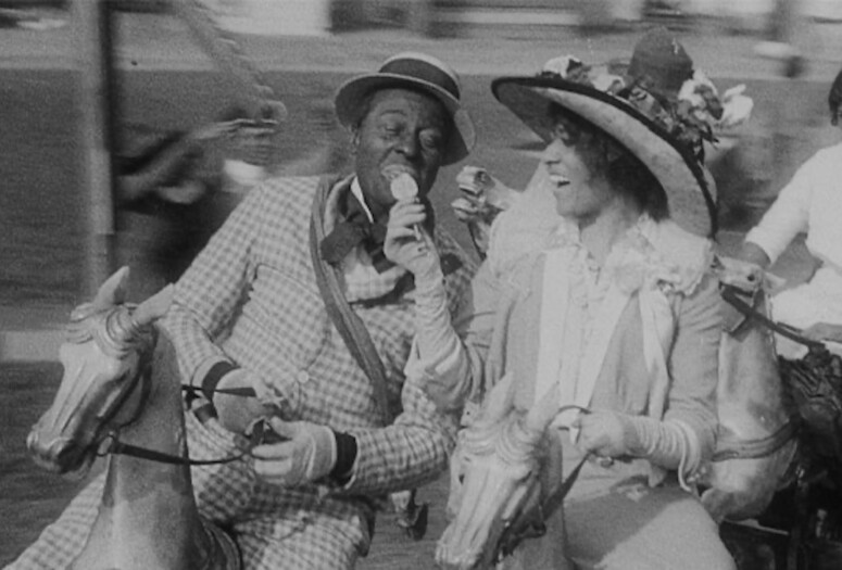 A still from the silent film Lime Kiln Club Field Day (1913), featured in Garrett Bradley’s America (2019). Image alt text: black-and-white still from the 1913 film Lime Kiln Club Field Day. Actor Bert Williams sits atop a horse on a carousel, to the left of actor Odessa Warren Grey. He is wearing Blackface, a plaid suit, and a small brimmed hat. She sits atop a carousel horse to the right of him, wearing a large brimmed hat decorated with flowers. She has an open-mouthed smile, looking at Williams and sharing her ice cream with her right hand.