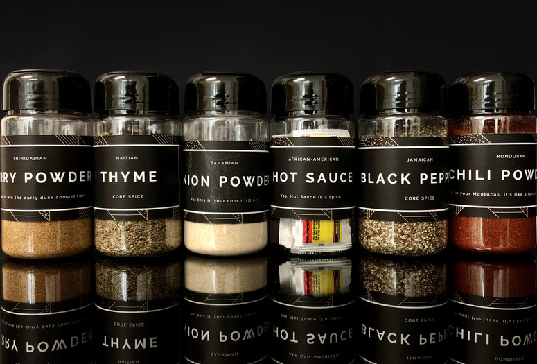 Image caption: Studio Barnes. Spices from A Spectrum of Blackness: The Search for Sedimentation in Miami. 2020. Plastic, spices, paper, and adhesive Image description: Photo of six bottles of spices against a black background and a reflective black surface. Each bottle has a black label with white text. The labels read from left to right: Trinidadian/ Curry Powder/ to win the curry duck competition; Hatian/ Thyme/ Core Spice; Bahamian/ Onion Powder/ Put this on your conch fritters; African-American/ Hot Sauce/ Yes, Hot Sauce is a spice; Jamacain/ Black Pepper/ Core Spice; Hondorun/ Chili Powder/ in your Montucas, it’s like tamale.