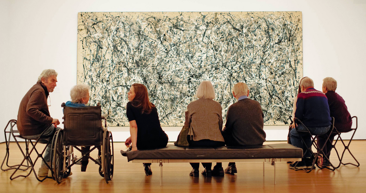 Algunos adultos mayores disfrutan de un Jackson Pollock durante uno de los Programas de acceso del MoMA. De “Para cuidadores: una guía para conectar de manera creativa”. Descripción de la imagen: En una galería, hay un grupo de siete adultos sentados de espaldas a la cámara, frente a un gran cuadro de arte abstracto. La mayoría tiene el cabello cano. Una mujer más joven de cabello largo castaño claro gira a su izquierda para hablar con uno de los adultos mayores que está en silla de ruedas. Al fondo, la pintura, tan grande que solo se ve parcialmente, presenta salpicaduras en arco de pintura negra, blanca y gris.