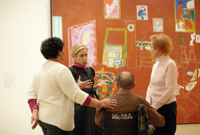 A photograph of an educator in conversation with three adults in front of the large-scale painting The Red Studio by the artist Henri Matisse. Photo: Jason Brownrigg. Digital Image © 2022 The Museum of Modern Art, New York