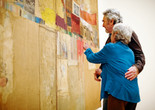 A photograph of two embracing adults looking closely at the large assemblage artwork Rebus by the artist Robert Rauschenberg. Photo: Jason Brownrigg. Digital image © 2022 The Museum of Modern Art, New York