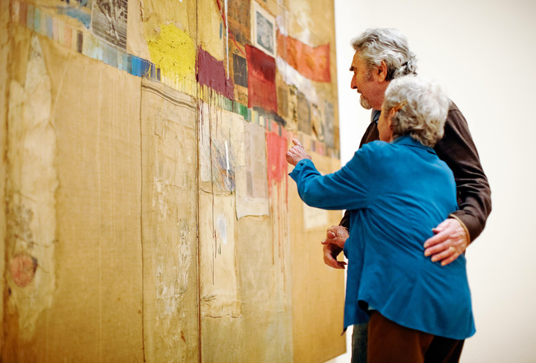 A photograph of two embracing adults looking closely at the large assemblage artwork Rebus by the artist Robert Rauschenberg. Photo: Jason Brownrigg. Digital image © 2022 The Museum of Modern Art, New York