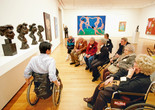 A photograph of an educator in conversation with a seated group of 10 adults facing a display of four sculpted bronze busts by the artist Henri Matisse in a gallery with colorful paintings on the wall. Photo: Jason Brownrigg. Digital image © 2022 The Museum of Modern Art, New York