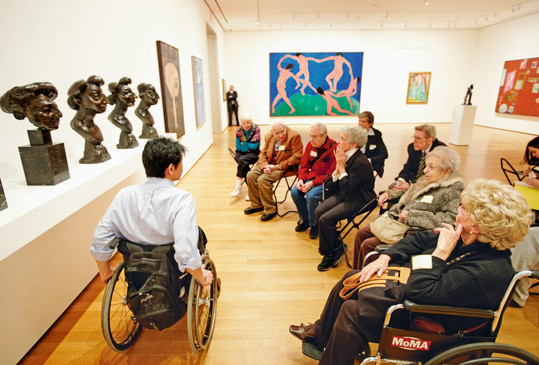 A photograph of an educator in conversation with a seated group of 10 adults facing a display of four sculpted bronze busts by the artist Henri Matisse in a gallery with colorful paintings on the wall. Photo: Jason Brownrigg. Digital image © 2022 The Museum of Modern Art, New York