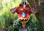 Lauren McArthur. Miss B. Is Kind but Gets a Little Shy. 2022. Multimedia. Photo: Mallory Perry Image description: A photograph of the artist wearing a colorful creature mask, with its tongue sticking out, rainbow beaded eyelashes, and big dangling wire earrings. She is cloaked in ivy and standing in front of a tree in Central Park on a sunny day.