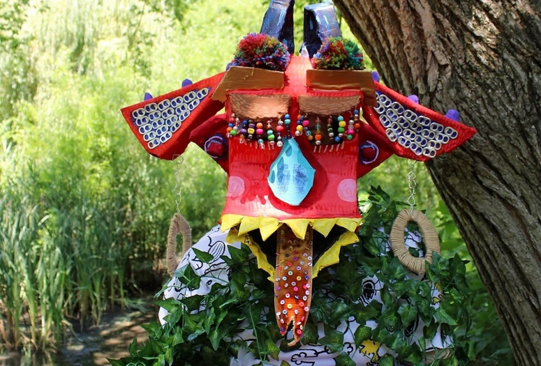 Lauren McArthur. Miss B. Is Kind but Gets a Little Shy. 2022. Multimedia. Photo: Mallory Perry Image description: A photograph of the artist wearing a colorful creature mask, with its tongue sticking out, rainbow beaded eyelashes, and big dangling wire earrings. She is cloaked in ivy and standing in front of a tree in Central Park on a sunny day.
