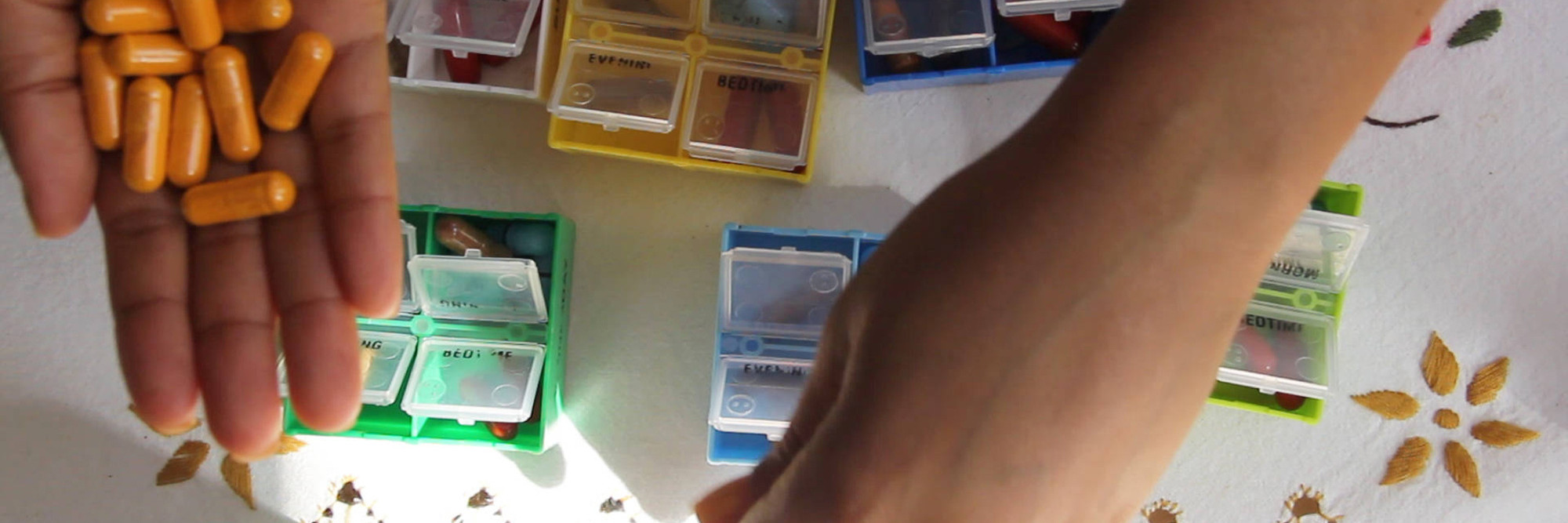 Carolyn Lazard. CRIP TIME. 2018. Video (color, sound), 10 min. The Museum of Modern Art, New York. Fund for the Twenty-First Century. © 2022 Carolyn Lazard Artist-provided description: In this video still, a set of hands sort seven pillbox compartments on an embroidered tablecloth. The image is taken from an overhead position with the hands partially obstructing the view of the differently colored compartments. The compartments are yellow, blue, pink, green, and white. They are filled with differently colored pills, too. There is a patch of bright sunlight on the tablecloth and there are a few pill bottles barely visible along the edge of the frame. Bright orange pills are held in the palm of one hand.