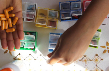 Carolyn Lazard. CRIP TIME. 2018. Video (color, sound), 10 min. The Museum of Modern Art, New York. Fund for the Twenty-First Century. © 2022 Carolyn Lazard Artist-provided description: In this video still, a set of hands sort seven pillbox compartments on an embroidered tablecloth. The image is taken from an overhead position with the hands partially obstructing the view of the differently colored compartments. The compartments are yellow, blue, pink, green, and white. They are filled with differently colored pills, too. There is a patch of bright sunlight on the tablecloth and there are a few pill bottles barely visible along the edge of the frame. Bright orange pills are held in the palm of one hand.