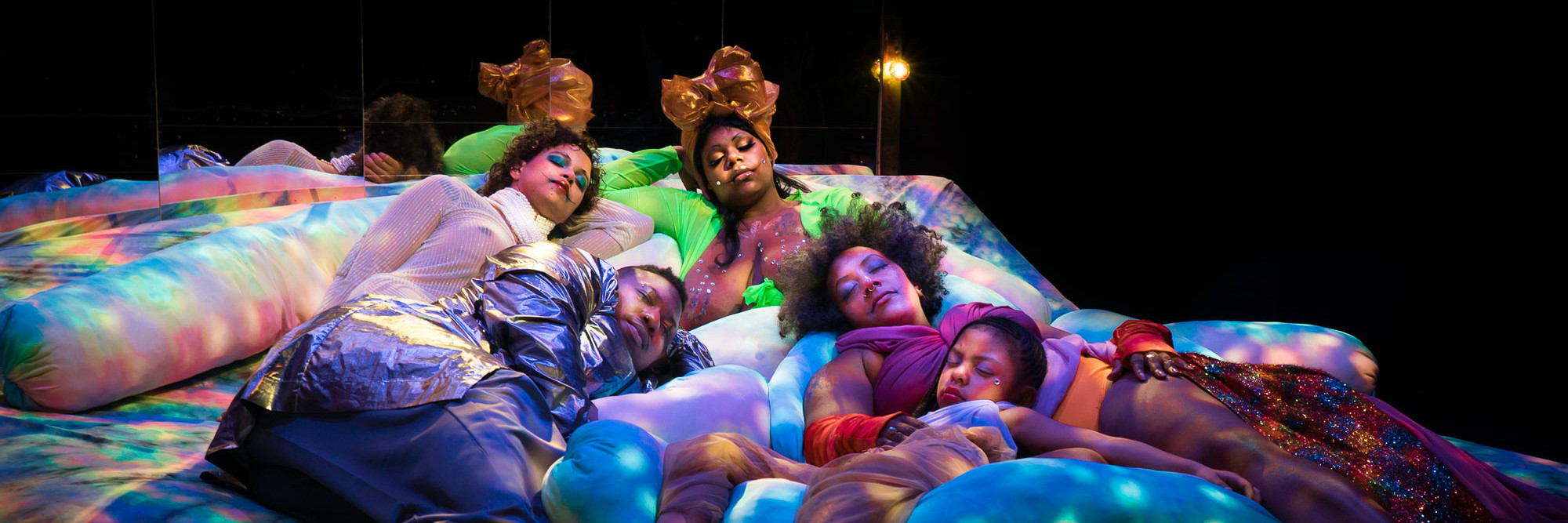 Black Power Naps installation at Performance Space, 2019. Courtesy of Black Power Naps. Photo: Avi Avion Image description: Four adults and one child sleep on colorfully dyed cushions under soft, dappled lighting.
