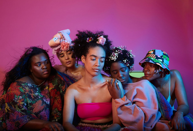 Black Power Naps installation. Photo by Xeno Rafaél Image description: Five beautifully dressed people wearing flowers in their hair sit close to each other, looking at each other with loving expressions.