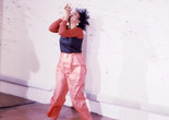 Senga Nengudi performing *Air Propo* at JAM, 1981. Courtesy Senga Nengudi Image description: A horizontally-oriented photograph shows the artist Senga Nengudi performing in the Just Above Midtown gallery. She wears pink drawstring pants and a navy and red long-sleeved top. Her hands are held to her lips to create an instrument, and she uses her breath to make sound.
