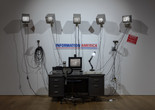Julia Scher. Information America. 1995. Metal office desk, five 9&#34; NTSC monitors with metal wall brackets, 13&#34; color monitor, plastic and vinyl signage, three black-and-white surveillance cameras, removable lenses, transformers, video matrix switchers, two time-lapse recorders, Amiga A1200HD computer, Sony Watch-Cam, two media players, desk lamp, office chair, wires and cables. The Museum of Modern Art, New York. The Jill and Peter Kraus Media and Performance Acquisition Fund. © 2023 Julia Scher