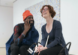 Photo: Martin Seck. ©️ 2023 The Museum of Modern Art, New York Image description: A photograph of dancer Jerron Herman and MoMA associate educator Annie Leist sitting together on black metal stools in a gallery. Jerron, a Black man wearing a neon orange beanie, holds his hand up to his face as he says something cheeky to Annie, a white woman wearing bright red glasses, who throws back her head in laughter.