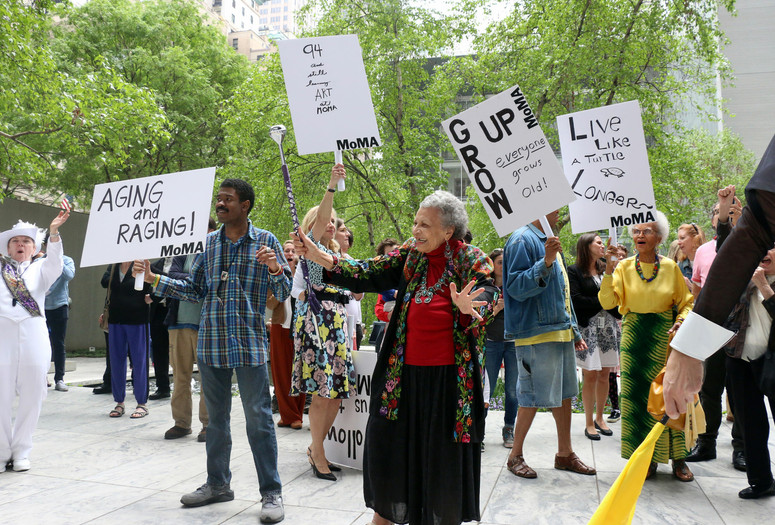 The Museum of Modern Art, New York. Photo: Manuel Molina Martagon Image description: A photograph of a group of older adults gathered outside in MoMA’s Sculpture Garden with a marching band. The crowd wears colorful clothing and hold signs that say “Aging and Raging!” and “Grow Up! Everyone Grows Old!”