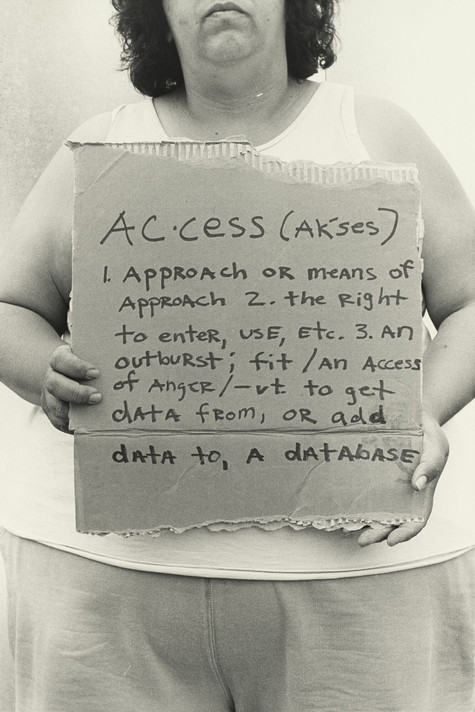 Laura Aguilar. Access + Opportunity = Success. 1993 Image description: A black-and-white photograph of the artist holding a cardboard sign with a paragraph of text. She wears a sleeveless white top and loose-fitting pants. The first line of the sign is the word “Access” followed by a phonetic pronunciation. The following lines are dictionary definitions of the term: “1. Approach or means of approach. 2. The right to enter, use, etc. 3. An outburst; fit/an access of anger/-vt. to get data from, or add data to, a database.” The photograph is cropped so that the top half of her head and her legs below the thigh are not included in the composition.