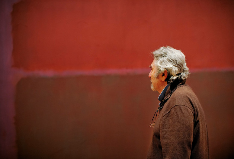 Photo: Jason Brownrigg ©️ 2024 The Museum of Modern Art, NY. Image description: Profile of a man with grey hair, a brown jacket, and reading glasses hung around his neck looking at a Rothko artwork with two red shaded squares on a lighter red background.