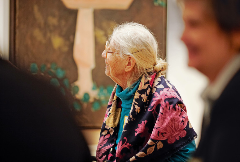 Photo: Jason Brownrigg ©️ 2023 The Museum of Modern Art, New York. Image description: Profile of a smiling woman with white hair and a purple floral scarf turned to look to the left in front of a brown and blue painting that fills the wall behind her.