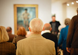 Photo: Jason Brownrigg ©️ 2024 The Museum of Modern Art, NY. Image description: A man in a tan corduroy jacket is centered among a blurred group of adults sitting with their backs turned to look at a framed artwork on the wall.