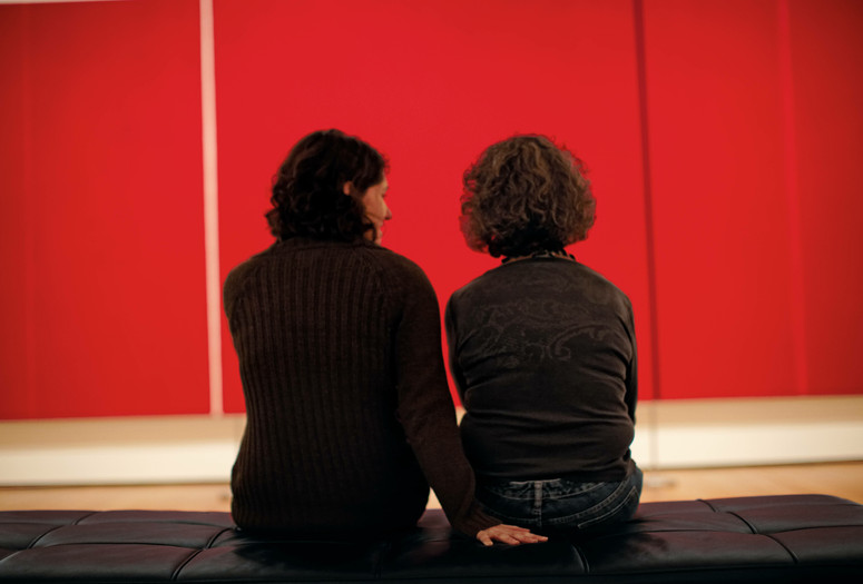 Photo: Jason Brownrigg ©️ 2024 The Museum of Modern Art, NY. Image description: Two women sit with their backs turned to the viewer, looking at a red canvas with vertical lines that hangs the whole span of the wall.