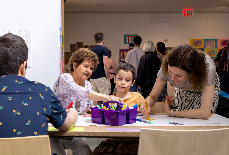 Photo: onwhitewall.com. © 2024 The Museum of Modern Art, NY Image Description: An older woman, a young boy, and a woman draw together at a long table covered in paper with a purple box of markers in a crowded space.