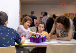 Photo: onwhitewall.com. © 2024 The Museum of Modern Art, NY Image Description: An older woman, a young boy, and a woman draw together at a long table covered in paper with a purple box of markers in a crowded space.