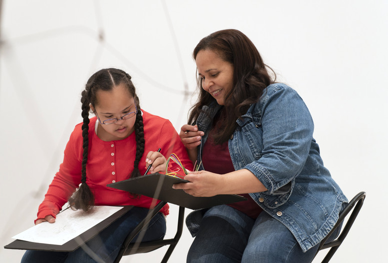 Photo: Martin Seck. © 2024 The Museum of Modern Art, NY Image Description: A girl in a red shirt and two braids intently draws on a black board that her mother wearing a jean jacket holds out to her.