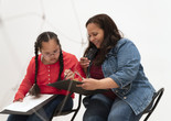 Photo: Martin Seck. © 2024 The Museum of Modern Art, NY Image Description: A girl in a red shirt and two braids intently draws on a black board that her mother wearing a jean jacket holds out to her.